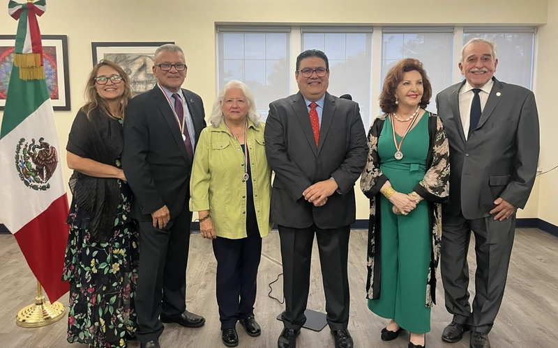 Recipients and officials pose together with their Ohtli Award medals at a presentation event next to a Mexican flag.