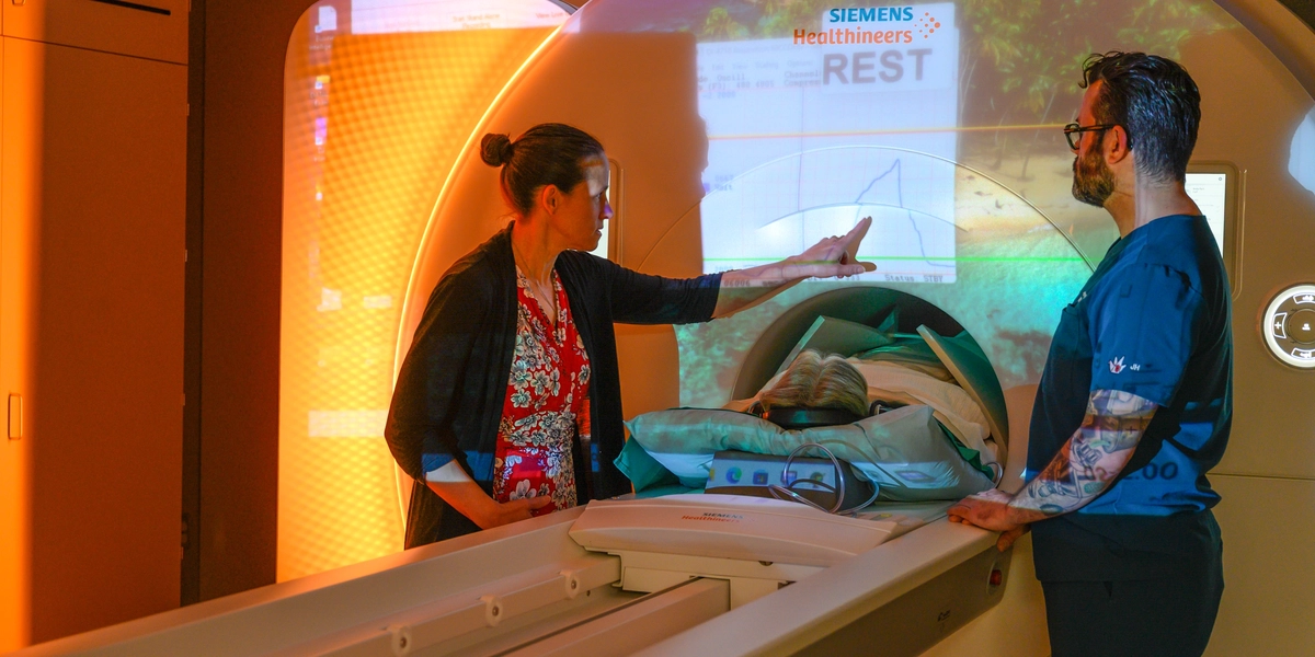 Two researchers stand beside a patient positioned inside MSU’s Biograph One imaging scanner as one points to projected medical data on the machine.