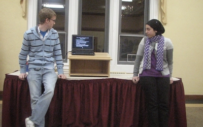 Nada and Ryan lean on a long table with a computer sitting on a wooden box.