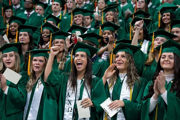 Graduates in green gowns cheer during an indoor commencement ceremony.