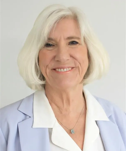 A photo of a woman with a short white blonde bob. She is wearing a lavender blazer over a white button down shirt against a light gray background.