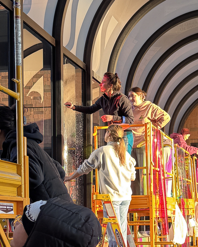 A group of students and faculty stand on scaffolding to install translucent film along glass in a glass, arched walkway