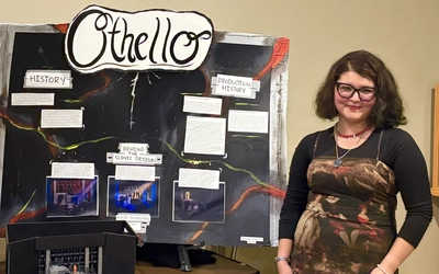 Ashley Romo poses in front of a presentation board that reads "Othello" with information about the Shakespeare play
