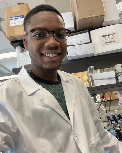 An undergraduate student wearing a white lab coat smiles for a selfie in a lab