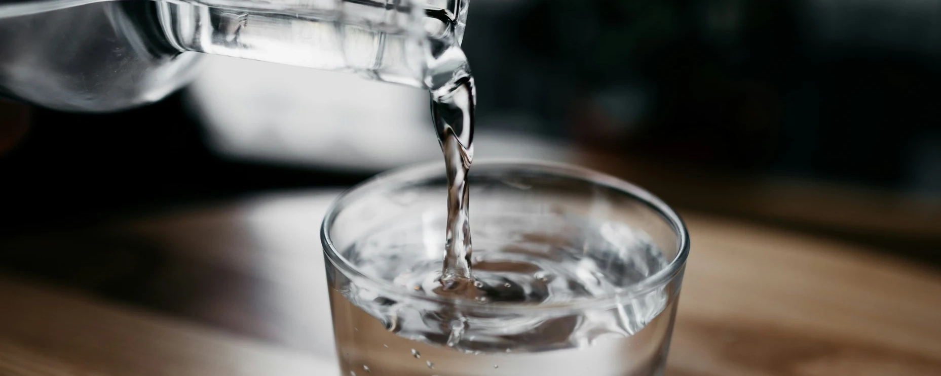 Clear glass bottle pours water into a glass on a wood table.