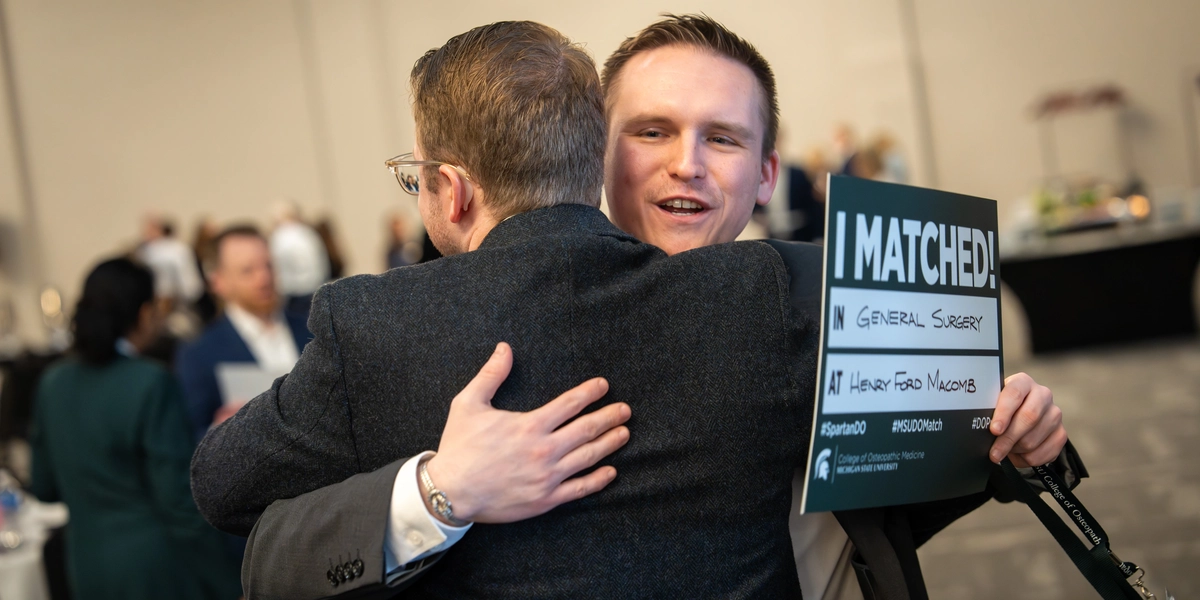 Evan Schumacher hugging a classmate while holding a "General Surgery" residency match sign.
