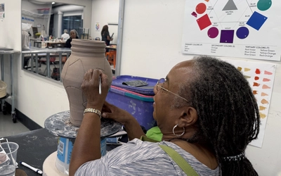 Valerie Horton draws a geometric design on a tall, unfinished clay vessel while working at a table in a Flint Institute of Arts studio.