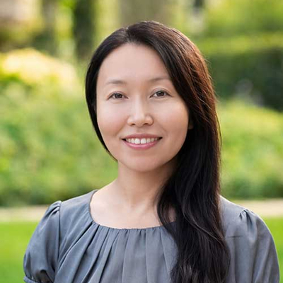 Portrait of Michigan State University sociology professor Xuefei Ren smiling outdoors, wearing a gray short-sleeved blouse, with greenery and a softly blurred campus landscape in the background.