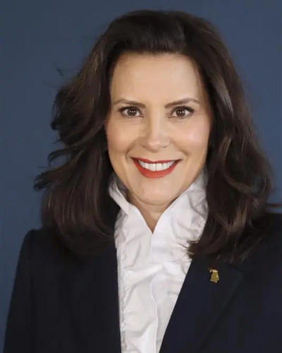 A picture of a woman with brown curled hair, wearing a black blazer, a white ruffled shirt and a gold Michigan shaped pin.