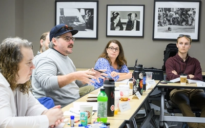 A man wearing glasses, a grey sweatshirt, and a baseball cap gestures while speaking during a group discussion around a meeting table with other participants listening.