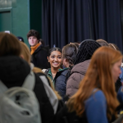 A candid shot of a smiling student with dark hair pulled back, surrounded by a dense crowd of peers in a hallway.