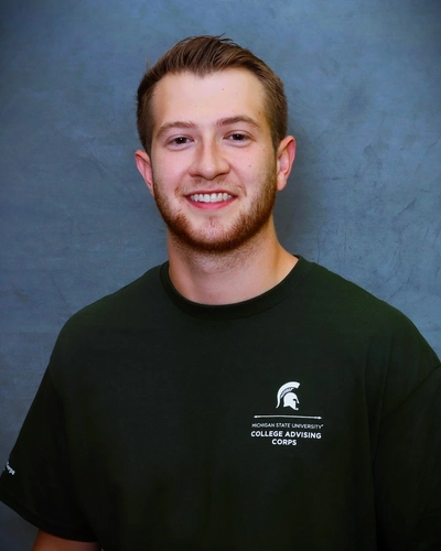 A headshot of a smiling young man with short brown hair and a light beard. He is wearing a dark green t-shirt that features the Michigan State University College Advising Corps logo on the chest. The background is a solid, textured grey.