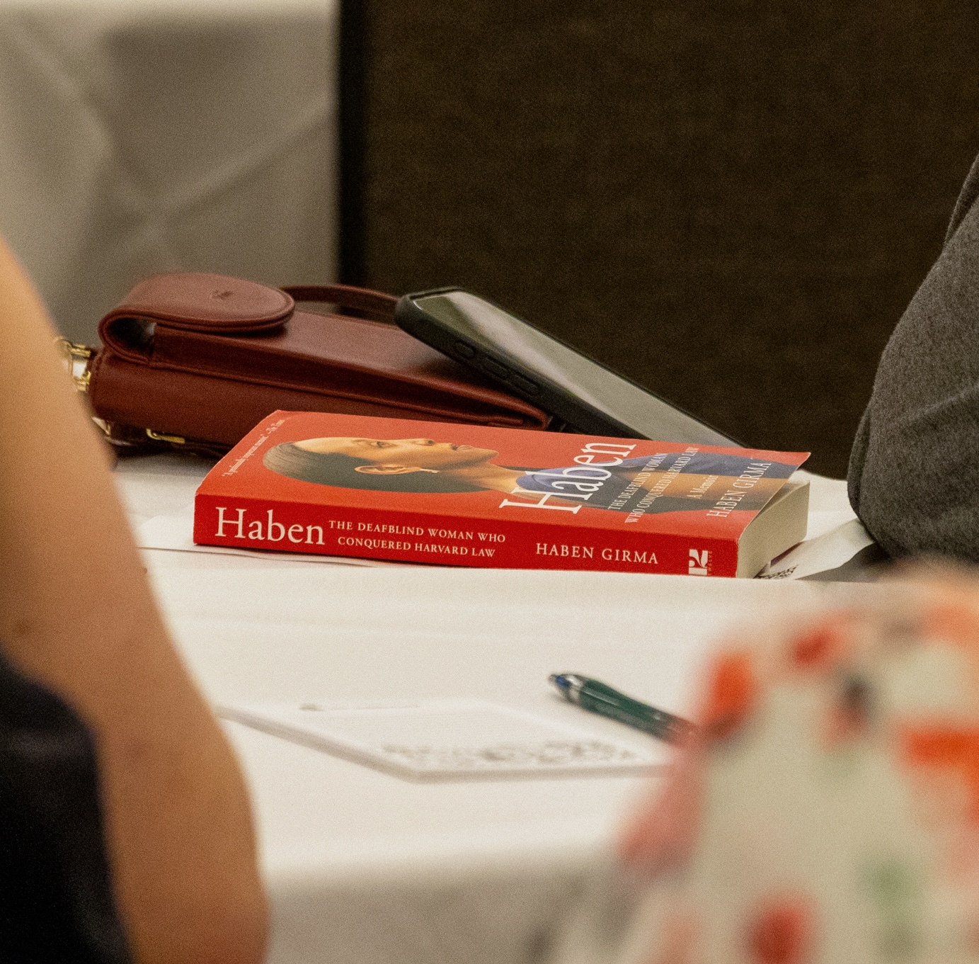 Haben Girma's red paperback book "Haben The DeafBlind Woman Who Conquered Harvard Law" sits on a conference table between attendees, a note pad and pen, and personal items. The book cover features Haben's image.