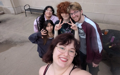 Five MSU students smile for an outdoor group selfie, with three holding up peace signs.