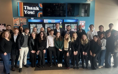 A group poses for a photo at a bar, above them a TV sign reads "THANK YOU"