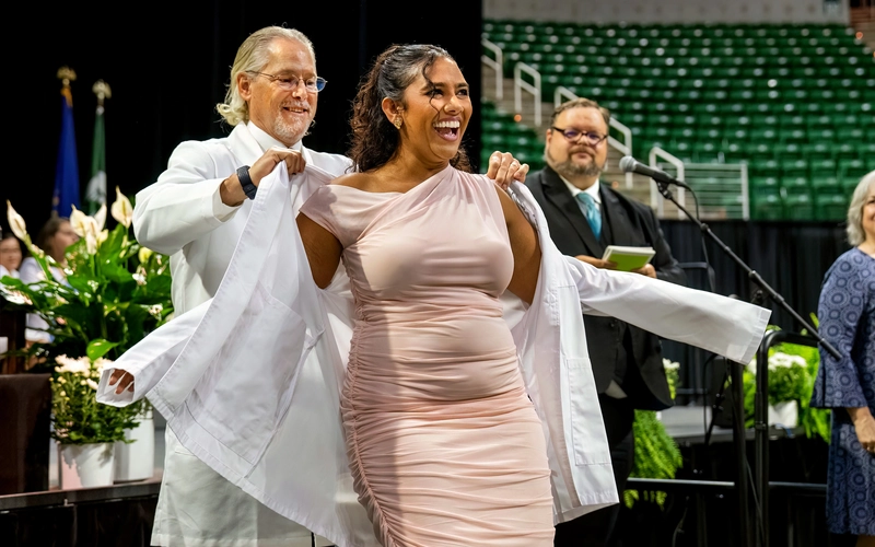 A smiling medical student is helped into their white coat by a faculty member on stage during the MSU College of Osteopathic Medicine White Coat Ceremony at the Breslin Center.