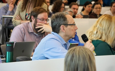 A close-up shot of an audience member with short graying hair and glasses speaking into a handheld microphone during a presentation. He is wearing a light blue striped button-down shirt. Other attendees sit behind him in tiered lecture hall seating, including a man with a beard resting his hand on his face while looking at a laptop.