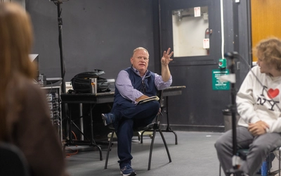Instructor seated with leg crossed gestures with left hand while facing group of students.