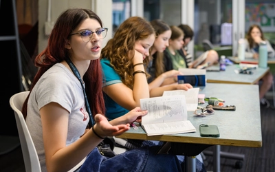 A student with reddish hair and glasses holds an open book while gesturing and speaking during a classroom reading discussion.