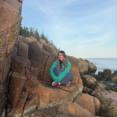A girl with blonde hair wearing a teal jacket and blue pants sitting on top of some rocks with a body of water in the background