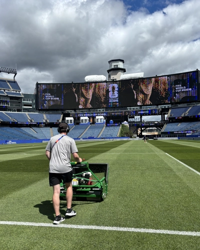 Elijah Delaney operates a walk-behind mower on a striped soccer pitch beneath a scoreboard displaying FIFA World Cup 2026 graphics.