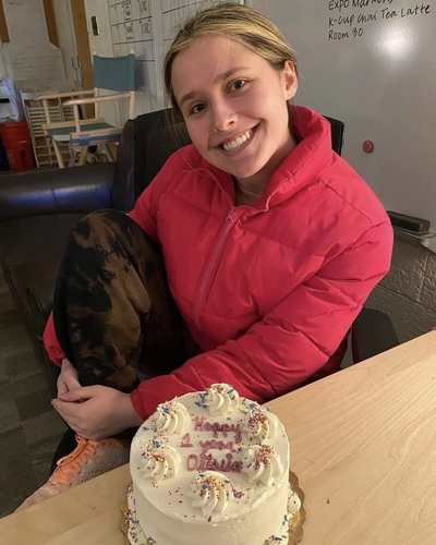 A girl with blonde hair wearing a red jacket sitting at a table. There is a white frosted cake in front of her that says "happy one year olivia"
