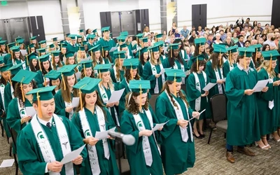 A large group of nursing students participating in an indoor ceremony for the Michigan State University College of Nursing. The graduates are dressed in green academic gowns with matching mortarboard caps and white stoles draped over their shoulders featuring the university logo. They stand in rows facing forward, reading from sheets of paper held in their hands as they recite their oath. An audience of family and friends is seated behind them in the background of the event space.