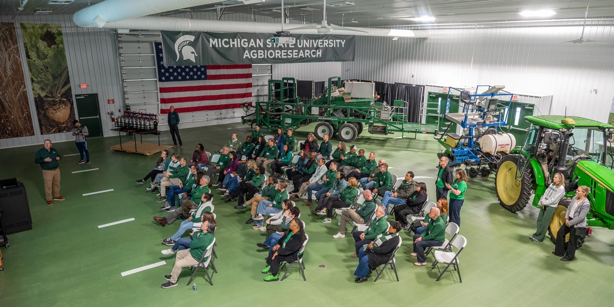 Group of people sit in chairs with a tractor behind them and a large banner that reads MICHIGAN STATE UNIVERSITY AGBIORESEARCH