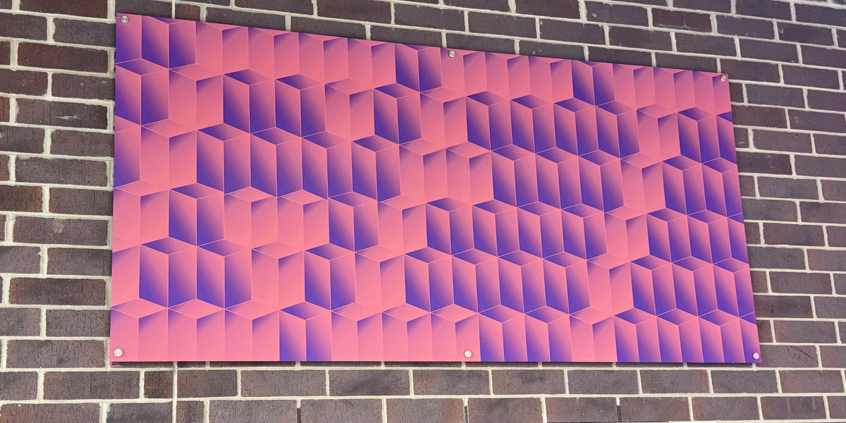 Abstract geometric wall artwork composed of repeating cube-like shapes in a pink-to-purple gradient, mounted on a brick wall above a window, with a small plaque identifying the piece nearby