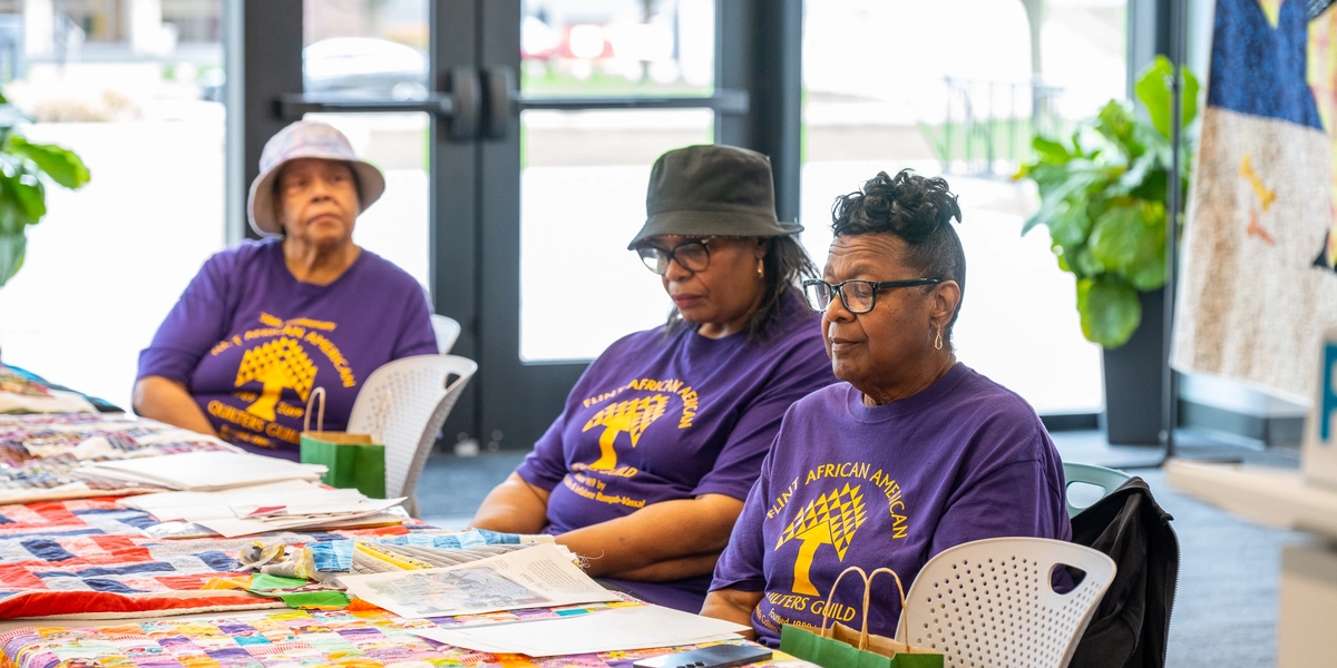 Three people in purple shirts sit at quilt-covered tables