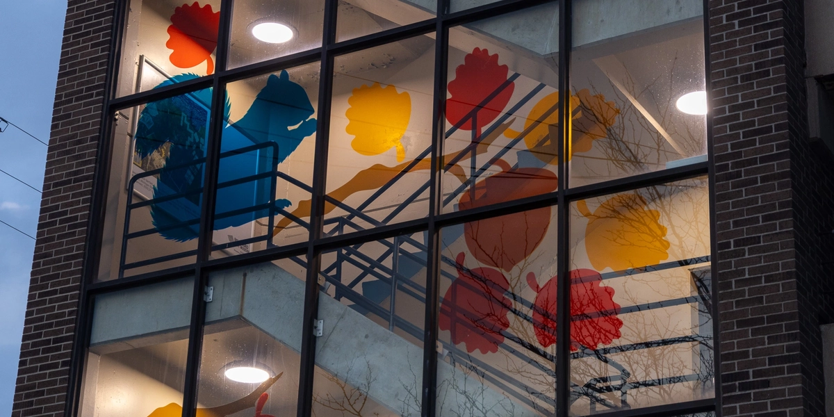Close-up of a glass-enclosed stairwell featuring colorful translucent floral shapes in blue, red, yellow, and orange, with stair railings and ceiling lights visible inside.