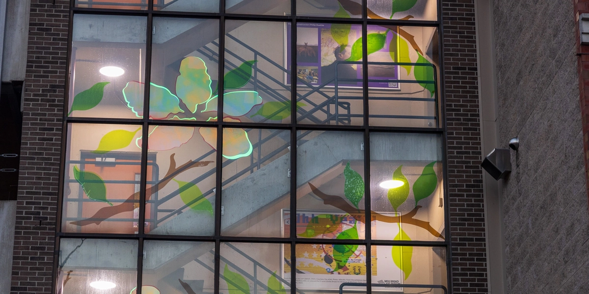Exterior view through a grid of windows revealing a stairwell decorated with large translucent green and yellow leaf and flower shapes applied to the glass