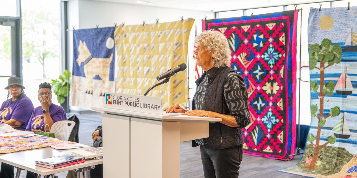 A person speaks at a podium in front of hanging quilts