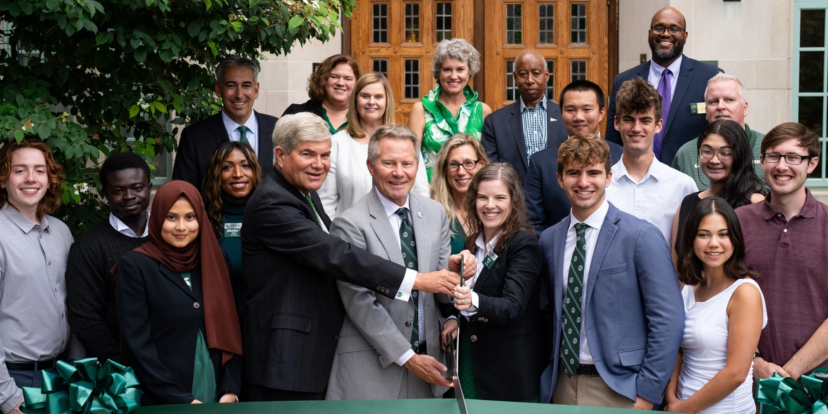 A large group of people, including students and MSU university leaders, stand smiling in front of a building with wooden double doors as they cut a green ribbon.