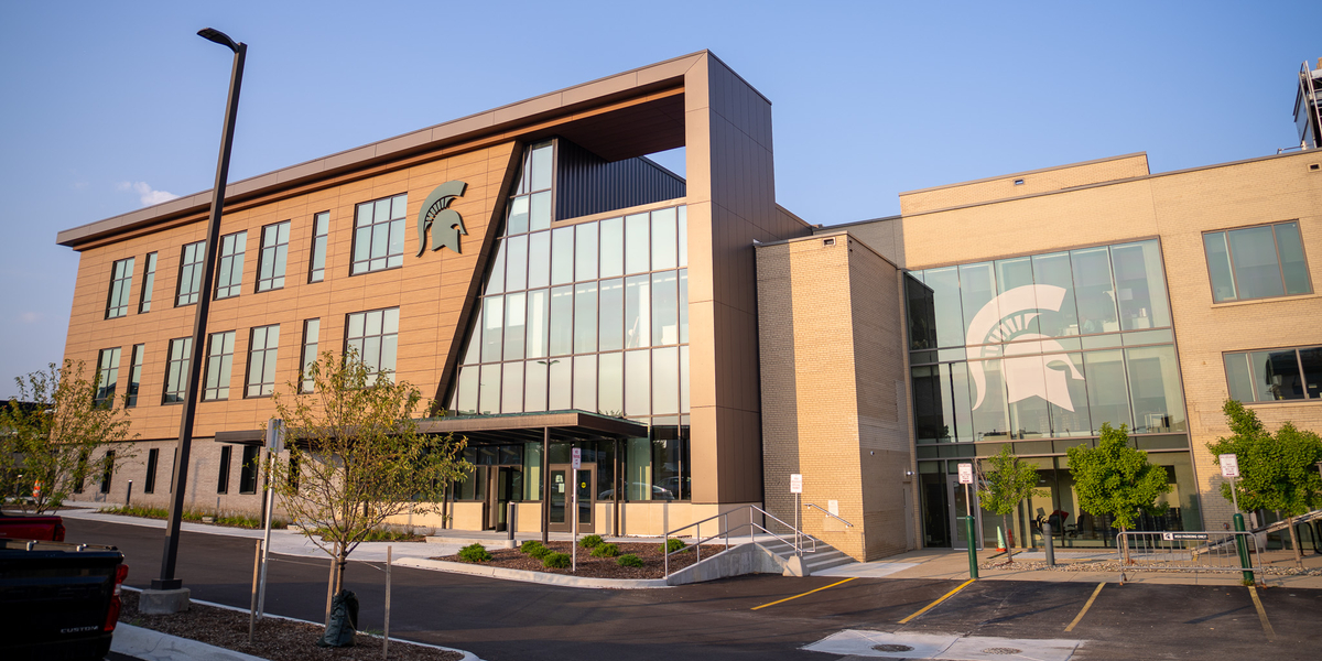 Exterior of the new Michigan State University College of Human Medicine public health facility in downtown Flint, a modern three-story building with glass walls and Spartan helmet logos.