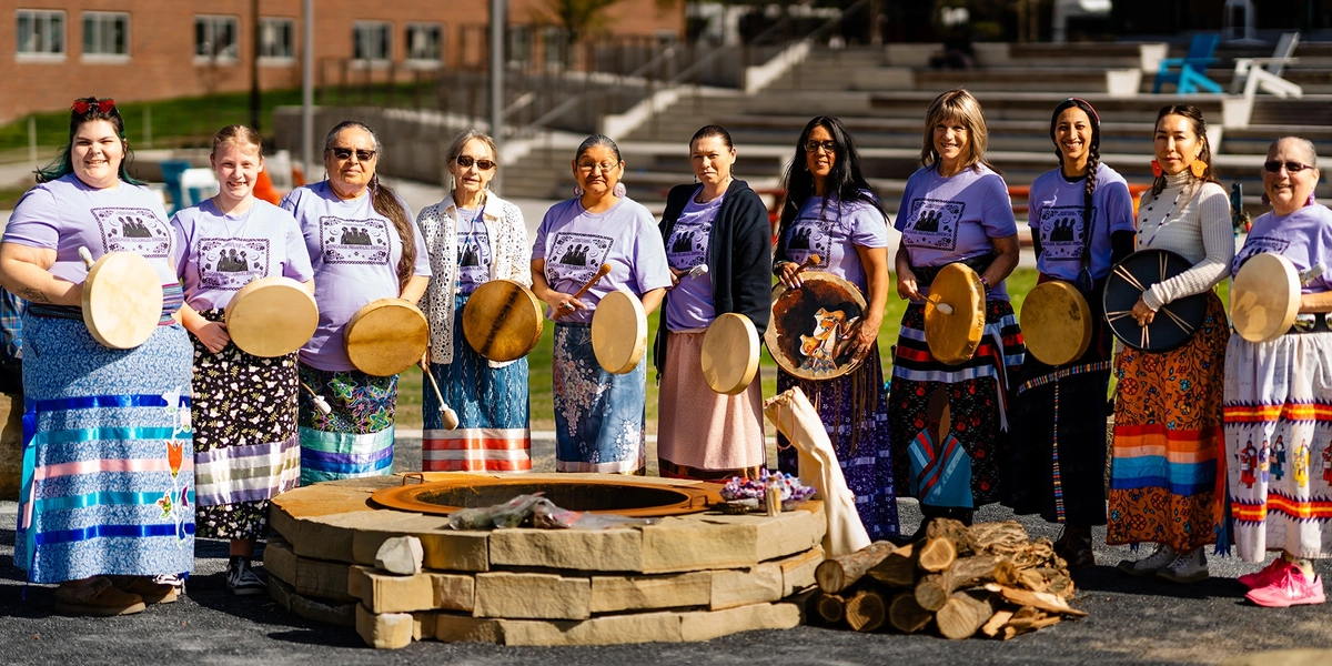 women drummers stand behind an outdoor fire pit