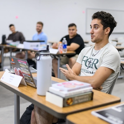 A photo of a young adult male in a cream colored tshirt sitting in a classroom. He has dark wavy hair and tan skin.