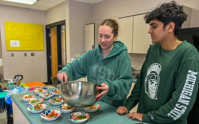 Two individuals stand at a counter in a classroom or kitchen setting, preparing food.