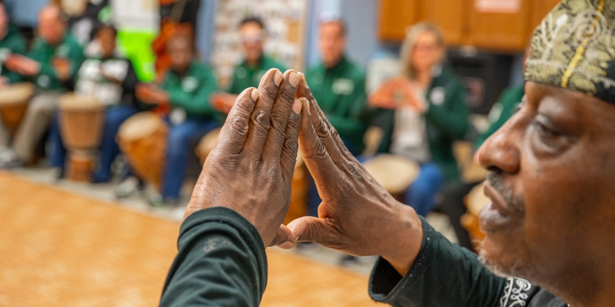Close-up photo of Kevin Collins, founder of the Kuungana African Drum and Dance Company, demonstrating a drumming technique with people sitting in the background
