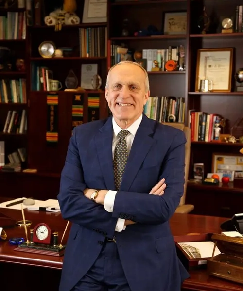 A photo of a man in a blue suit with a brown and tan spotted tie and white button down. He is leaning against a desk.
