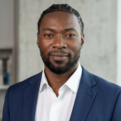 A professional headshot of a Black man with a neat beard and braided hair, looking directly at the camera with a subtle smile. He is wearing a tailored navy blue blazer over a crisp, white button-down shirt with an open collar, set against a soft-focus gray background.