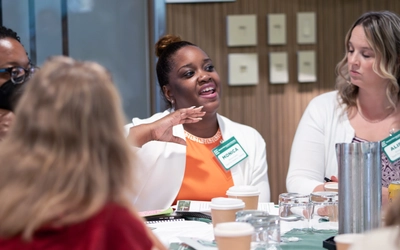 Participants engage in a small-group discussion around a table during a workshop, with one attendee gesturing while speaking as others listen. Coffee cups, notebooks, and name badges are visible on the table, highlighting the collaborative nature of the event.