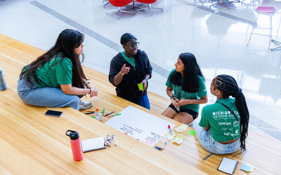 Four students in green T‑shirts sit on tiered wooden steps collaborating on a project, with markers, sticky notes, and a large poster board spread out between them in a bright indoor campus space.