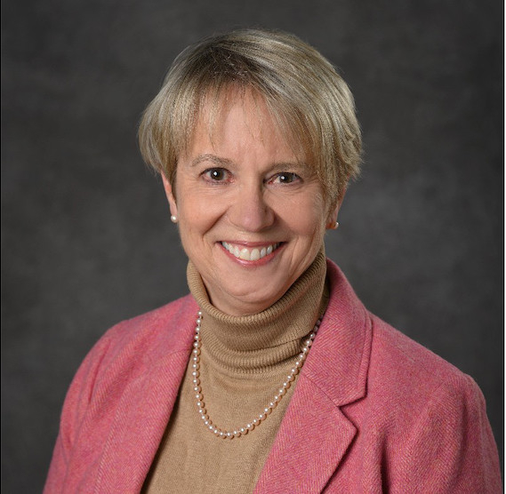 Professional headshot of Pamela Ruegg smiling at the camera, wearing a pink blazer over a tan turtleneck with a pearl necklace, against a dark gray studio background.