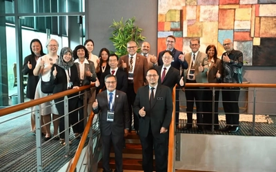 A group of professionally dressed adults wearing conference badges stand together on an indoor staircase, smiling and giving thumbs-up gestures. They are gathered in a modern building with glass walls, a large colorful abstract artwork, and a tall potted plant in the background.