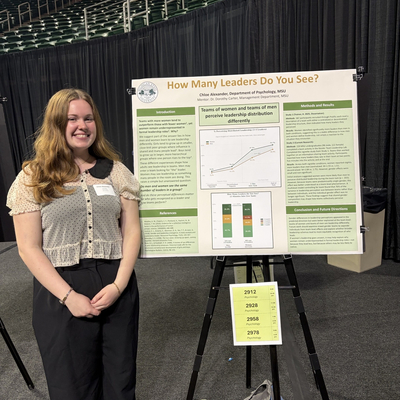A person stands smiling in front of an academic research poster titled “How Many Leaders Do You See?” The poster includes sections such as Introduction, Methods and Results, Conclusions and Future Directions, and References, along with a bar graph and a data table. The presenter wears a light-colored blouse and black pants. A black curtain backdrop, a display stand, and personal items like a water bottle and bag are visible around the setup.