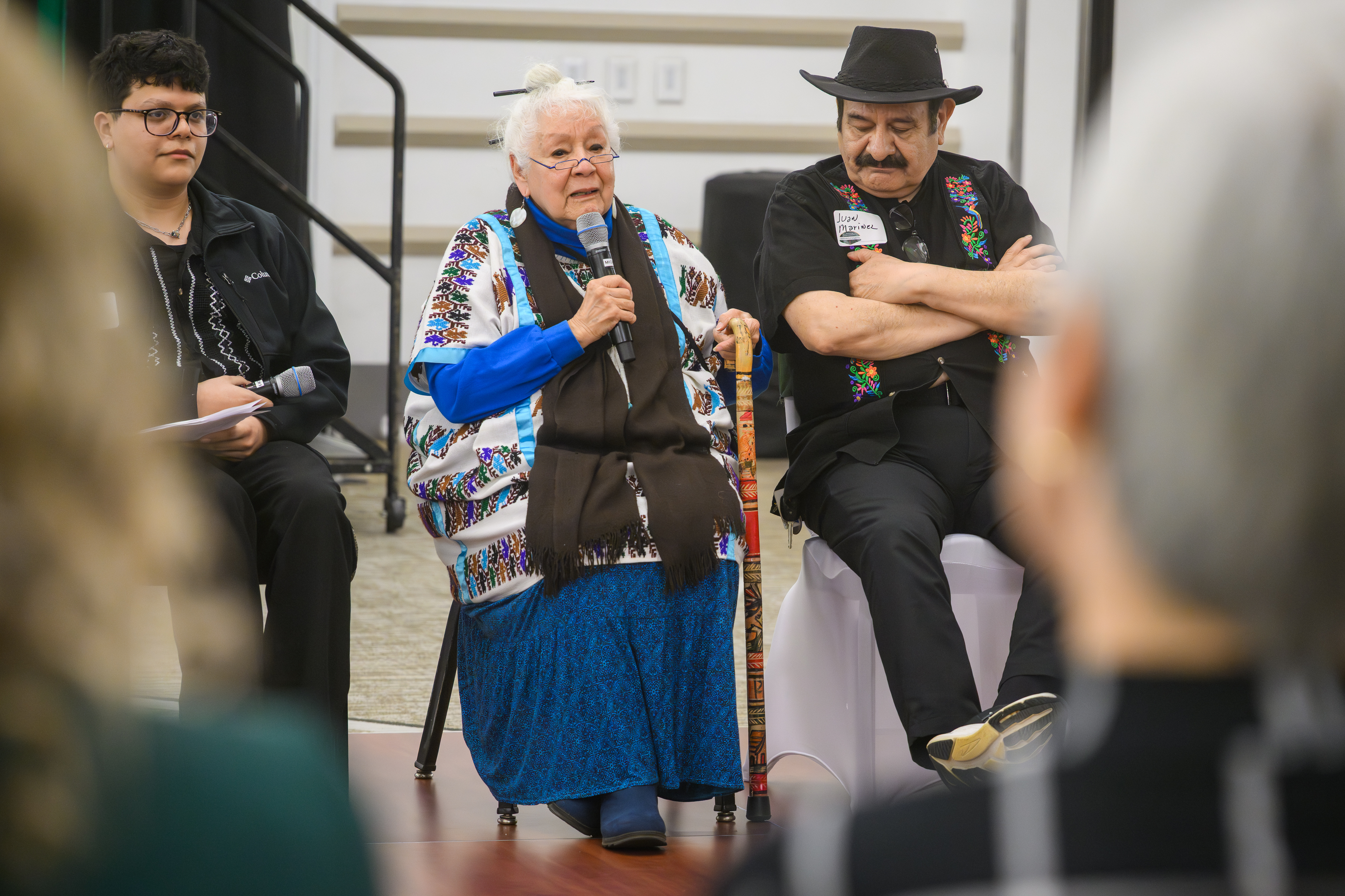 an elder woman speaks into a microphone while others listen