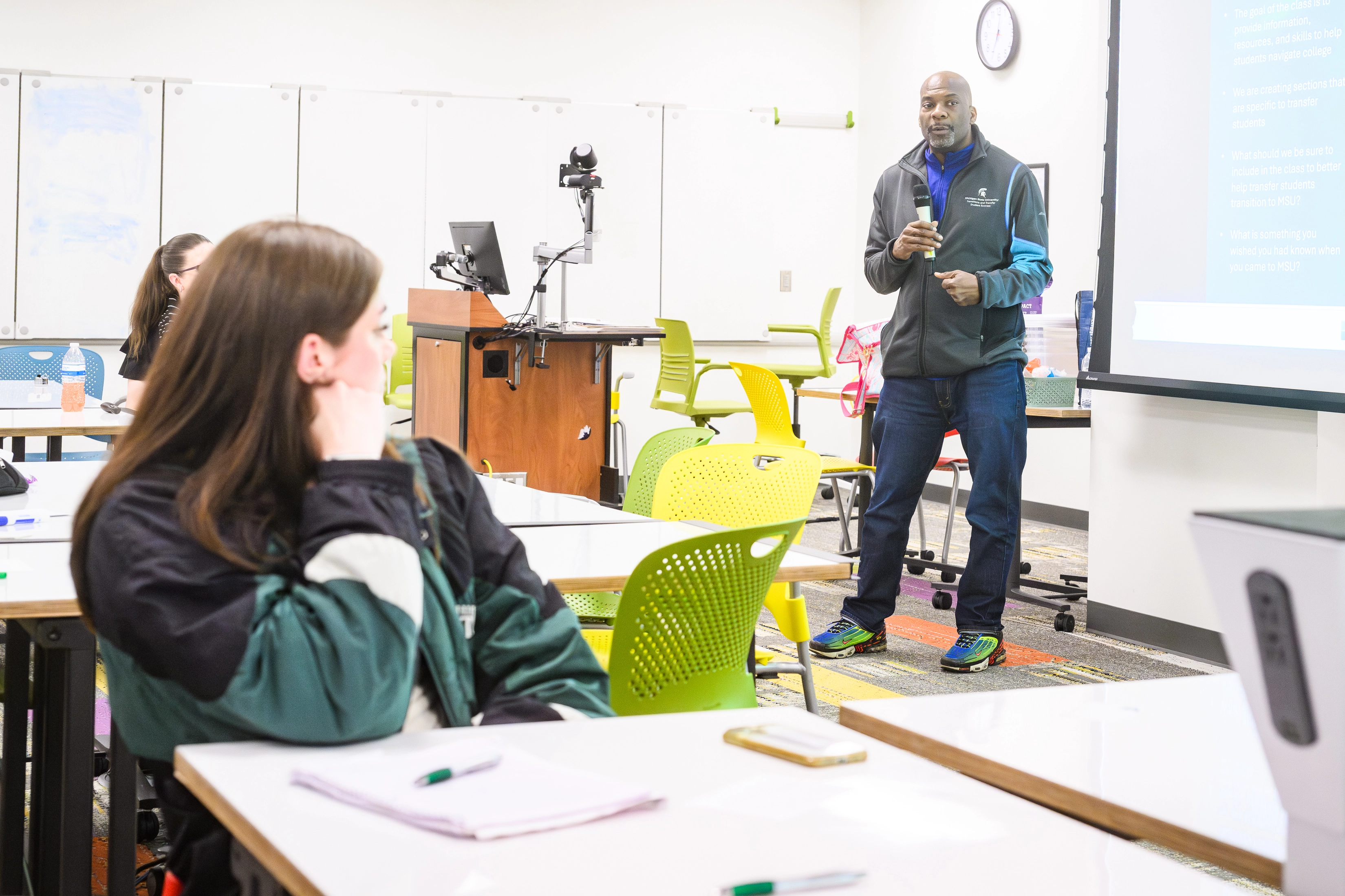 A man speaks into a mic, standing in a classroom in front of students