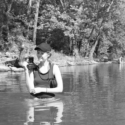 A person wearing a baseball cap stands waist-deep in a calm river while holding a camera. Trees line the riverbank in the background, and a small group of people can be seen farther downstream. The black-and-white photograph captures a fieldwork or photography scene in a natural outdoor setting.