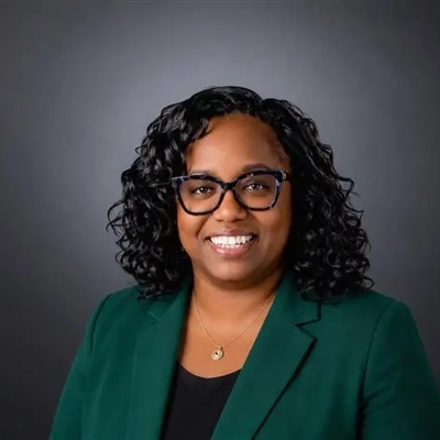 A photo of a woman with black glasses and curly black hair. She is wearing a green blazer with a black shirt underneath against a dark gray background.